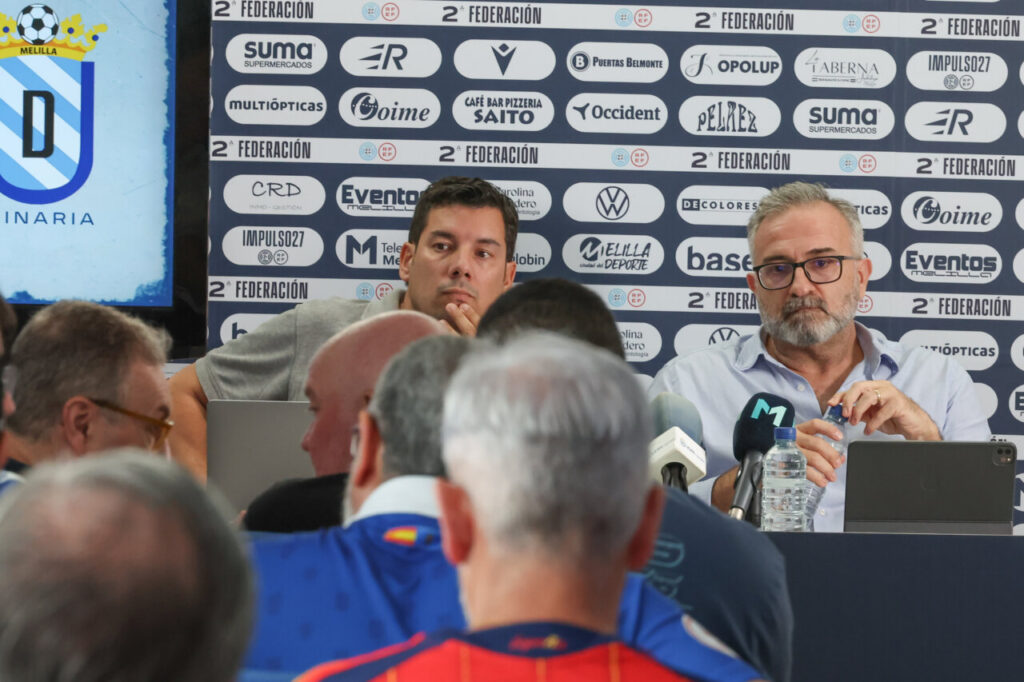 Press conference with two men at a table, multiple microphones, and a wall of sponsor logos behind them.