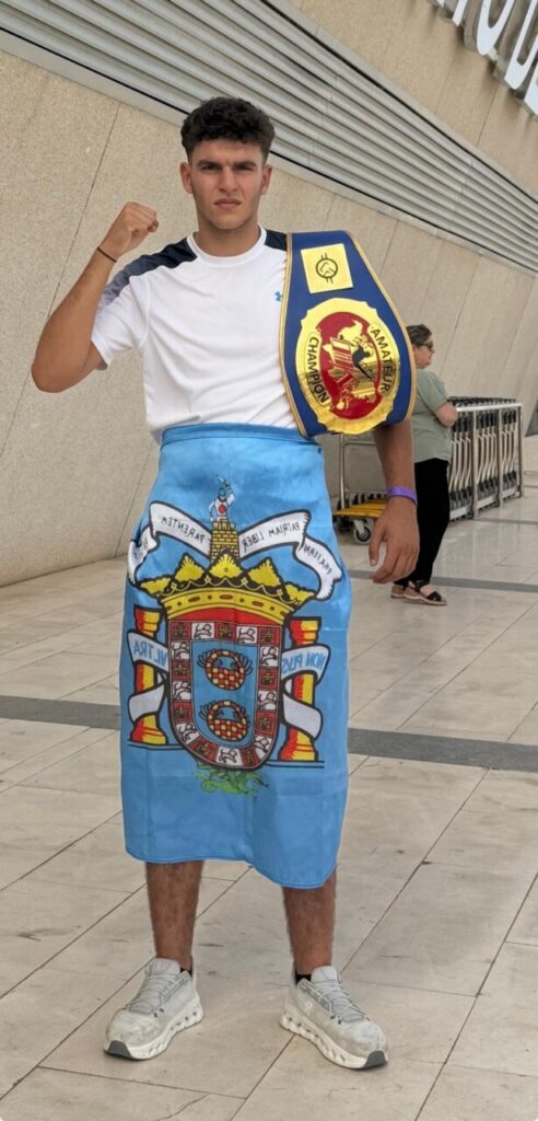 Young man indoors, wrapped in a blue towel featuring Portugal's coat of arms, posing with a championship belt over his shoulder and raising a fist.