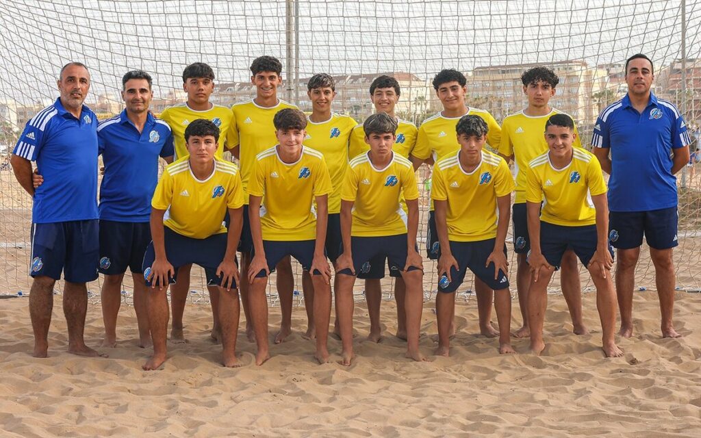 Youth soccer team in yellow jerseys and blue shorts posing on a sandy beach with a goal net behind them.