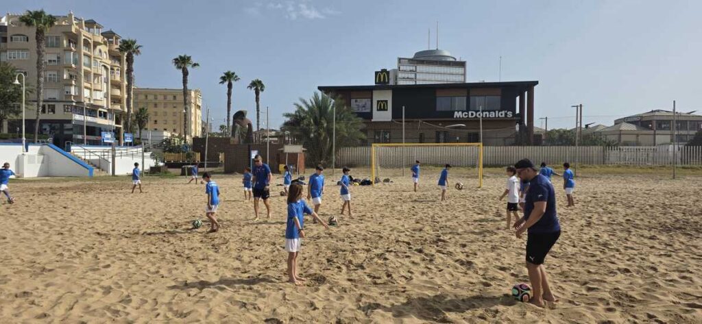 Niños entrenando fútbol en la playa con un fondo de edificios
