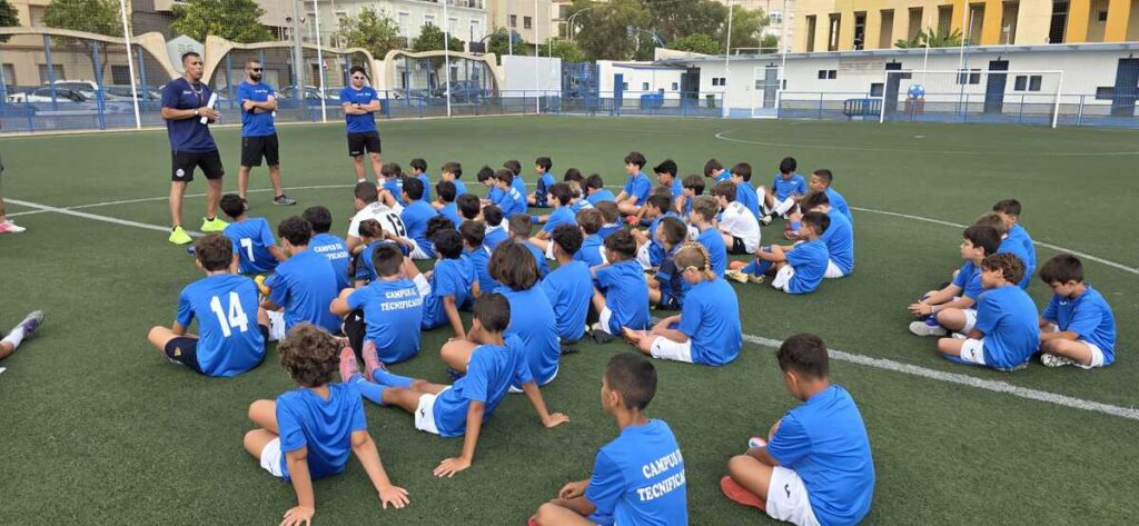 Young soccer players in blue uniforms sit on a turf field as coaches give instructions at a practice session.