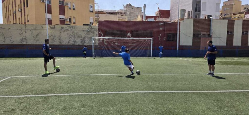 Young player in a blue shirt kicks a soccer ball toward the goal as teammates and a coach watch on an urban, fenced field.