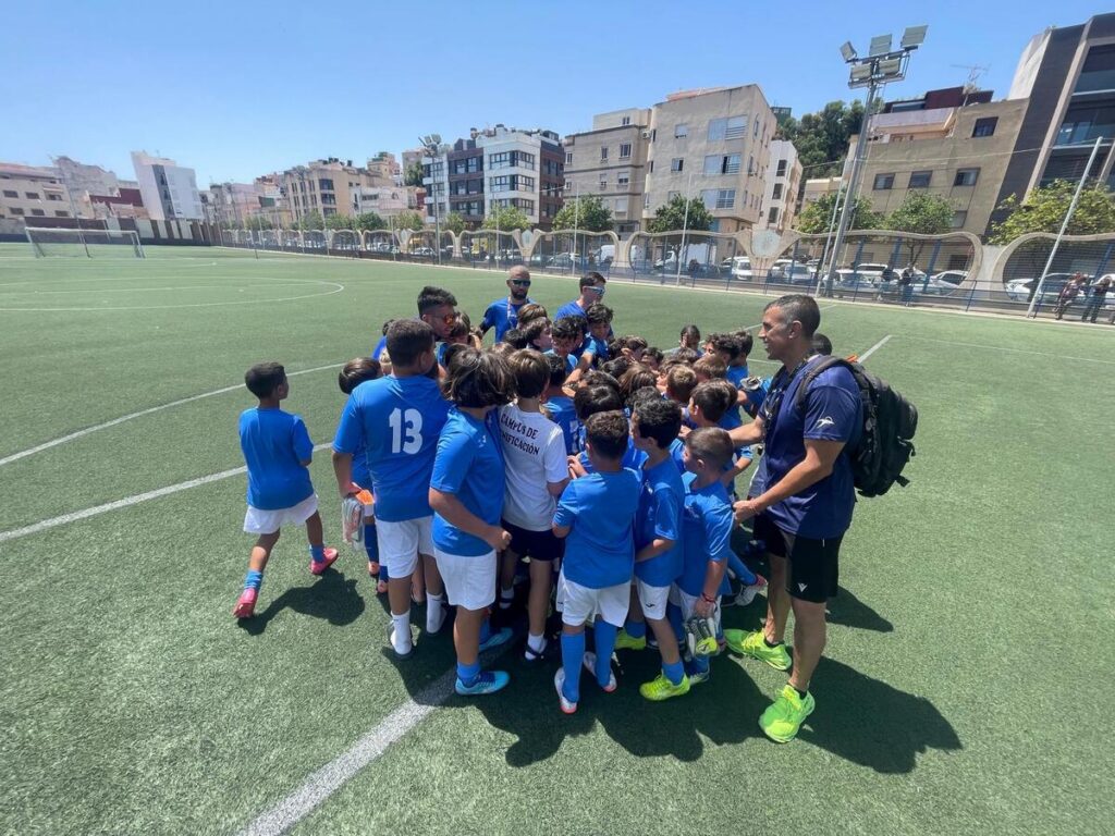 Group of young soccer players in blue uniforms huddle around coach on a sunny outdoor field.
