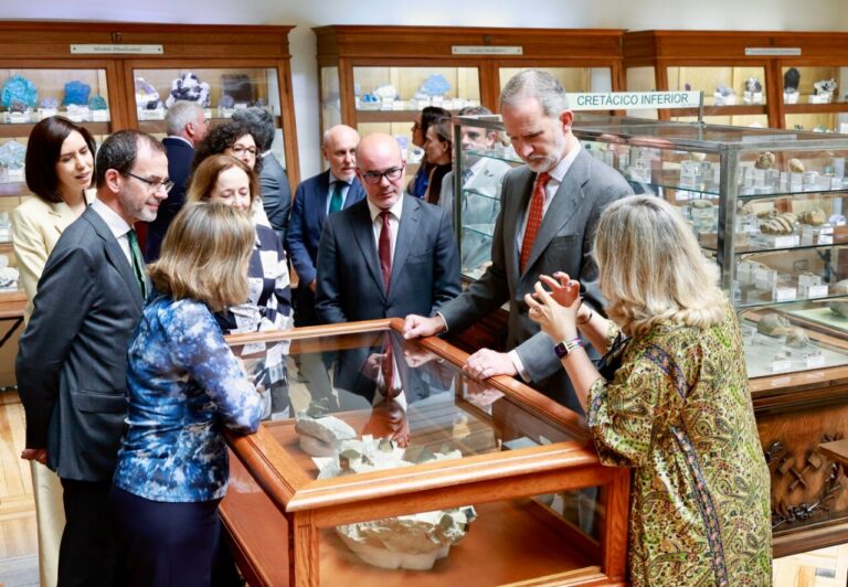 A group of professionals in suits gathers around a glass display case, examining mineral samples in a museum exhibit.