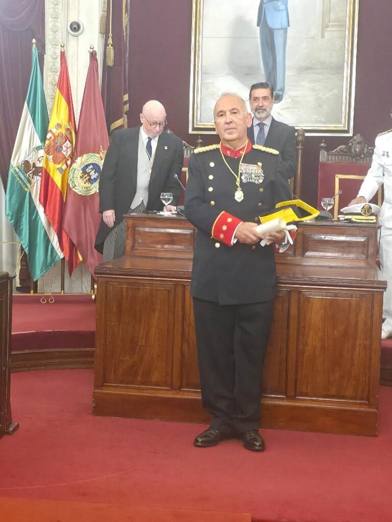Senior military officer in dress uniform with medals stands at a wooden podium during a formal ceremony, flags and officials in the background.