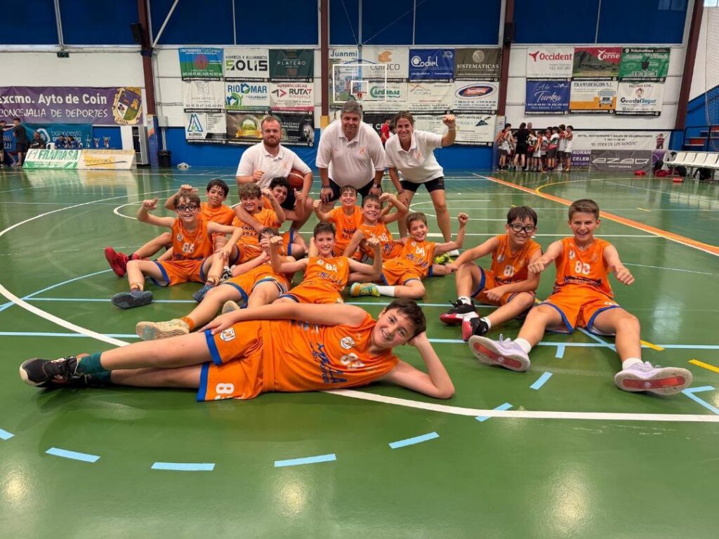 Group of young players in orange jerseys sit and lie on an indoor basketball court, posing with two coaches in white shirts behind them.