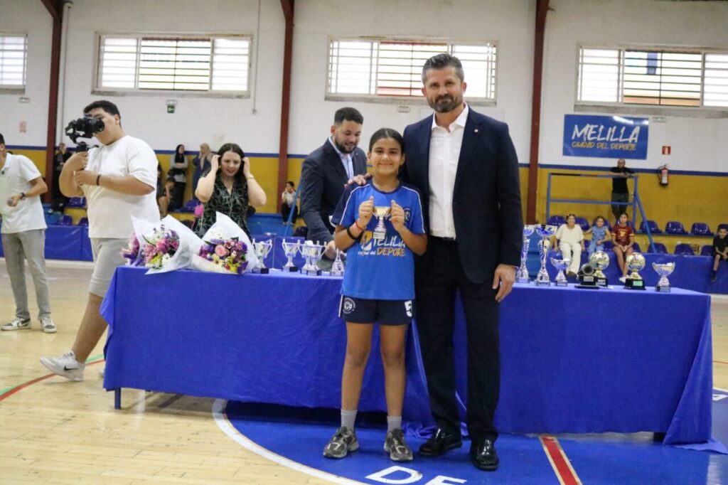 Young girl in blue sports kit standing with a man in a suit, posing with a trophy on a table of awards in a gymnasium.