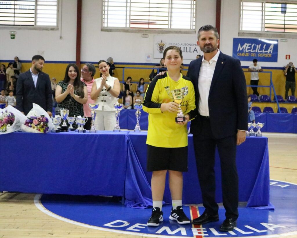 Young athlete in a yellow jersey holds a trophy beside a man in a suit during an awards ceremony in a sports hall.