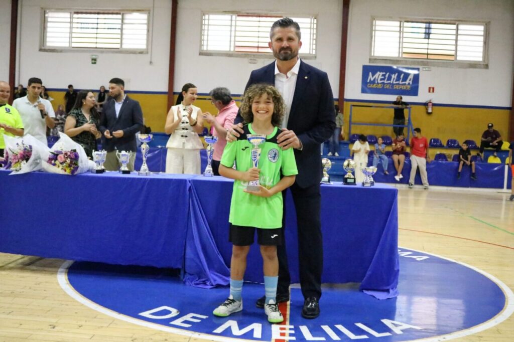 Boy in a green sports jersey holds a trophy, standing beside a man in a suit at an indoor awards ceremony.