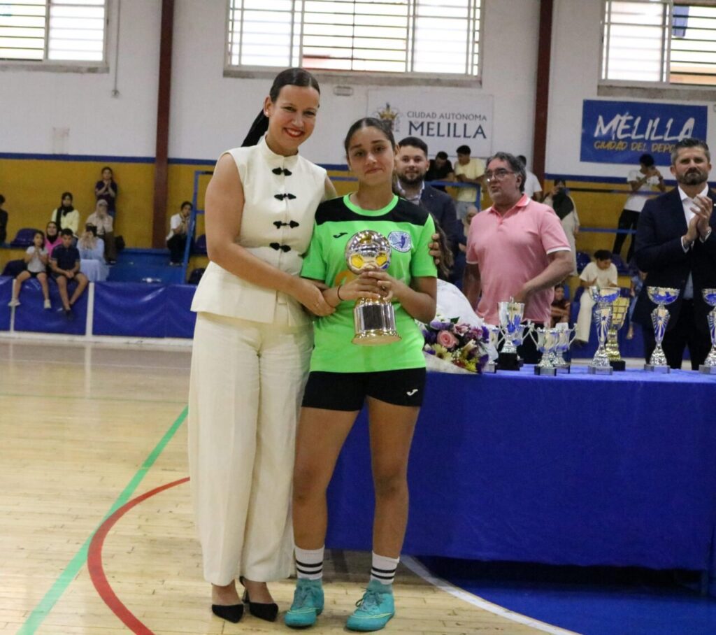 Woman in a white outfit stands with a young athlete in a neon green jersey, both smiling as the athlete holds a trophy at a gym awards ceremony.