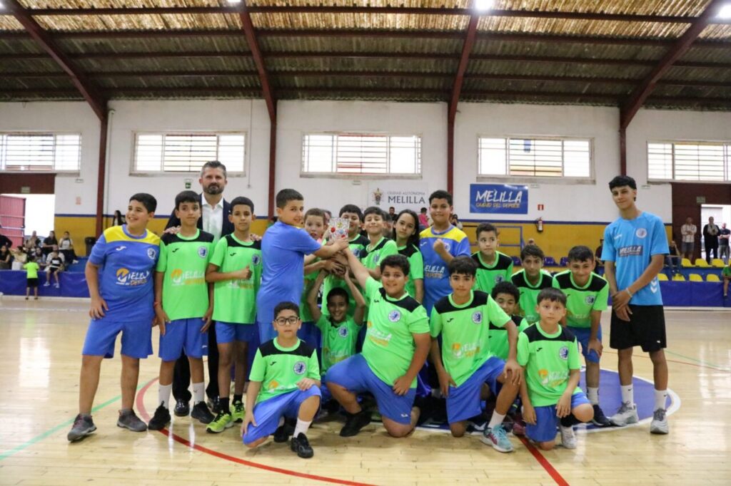 Group of boys in green and blue jerseys posing with a coach in an indoor gym hall.