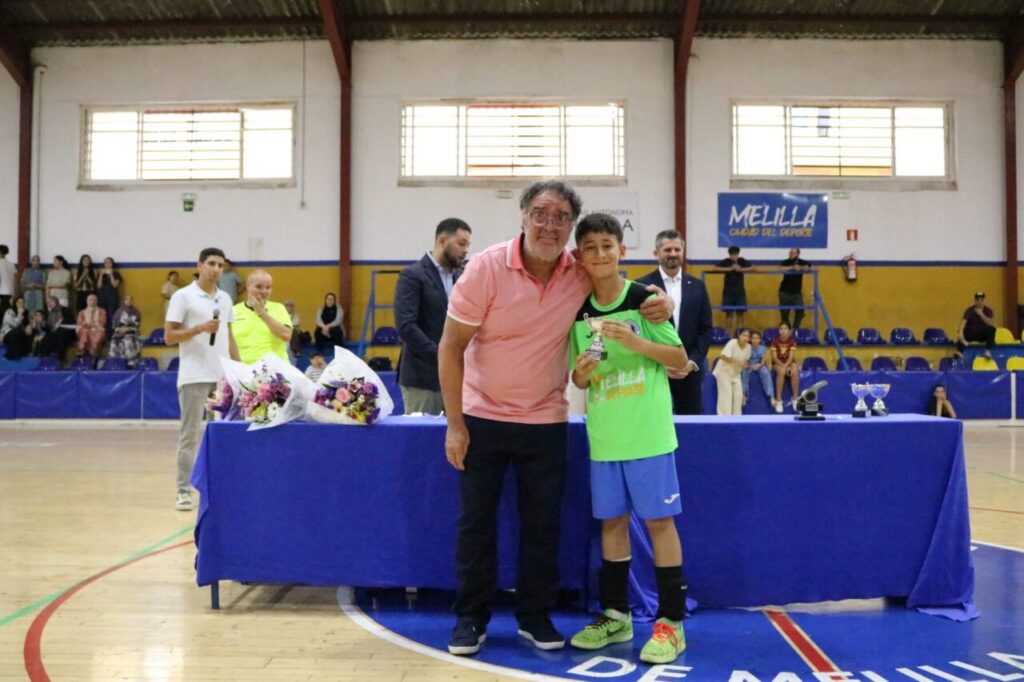 Man in pink polo hugs a young soccer player in a lime green shirt at an awards table in a gymnasium.