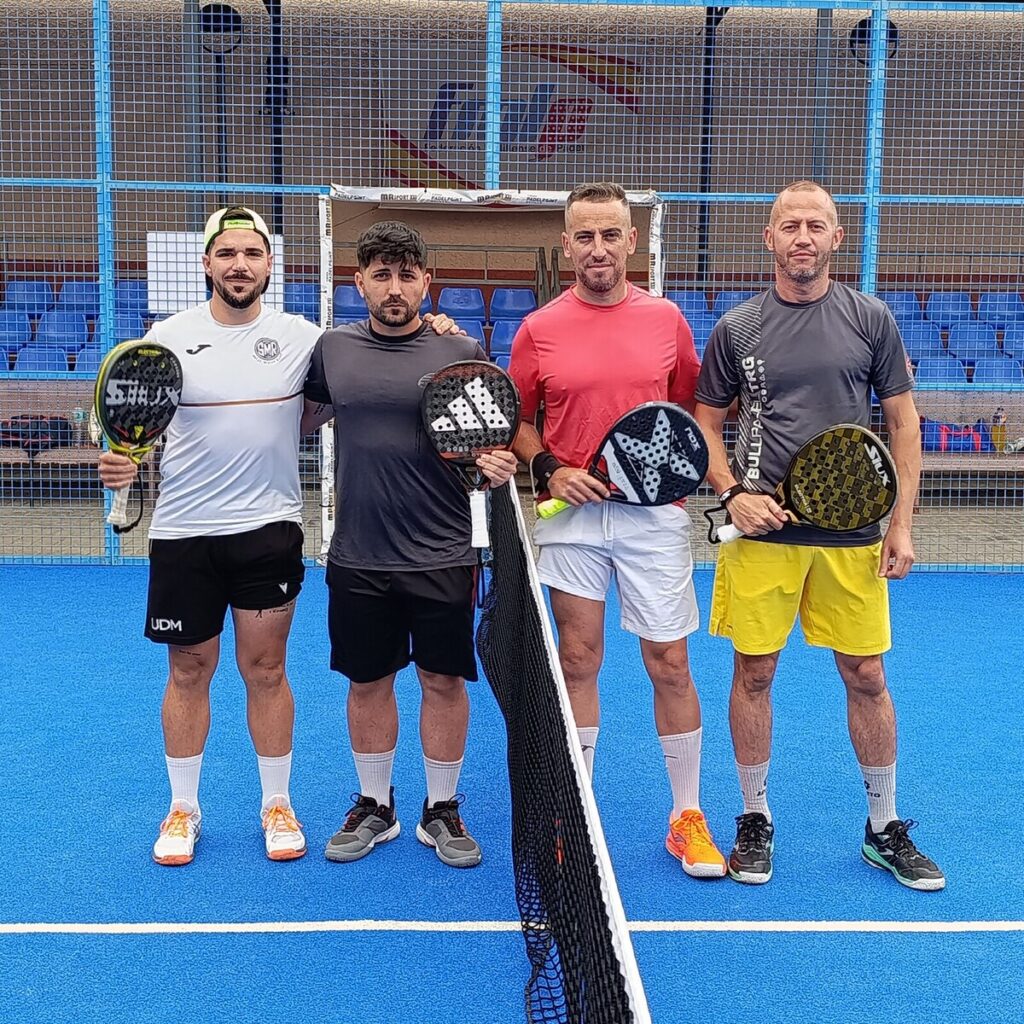 Four men posing with paddles on a blue padel court in front of a net and blue seating behind them.
