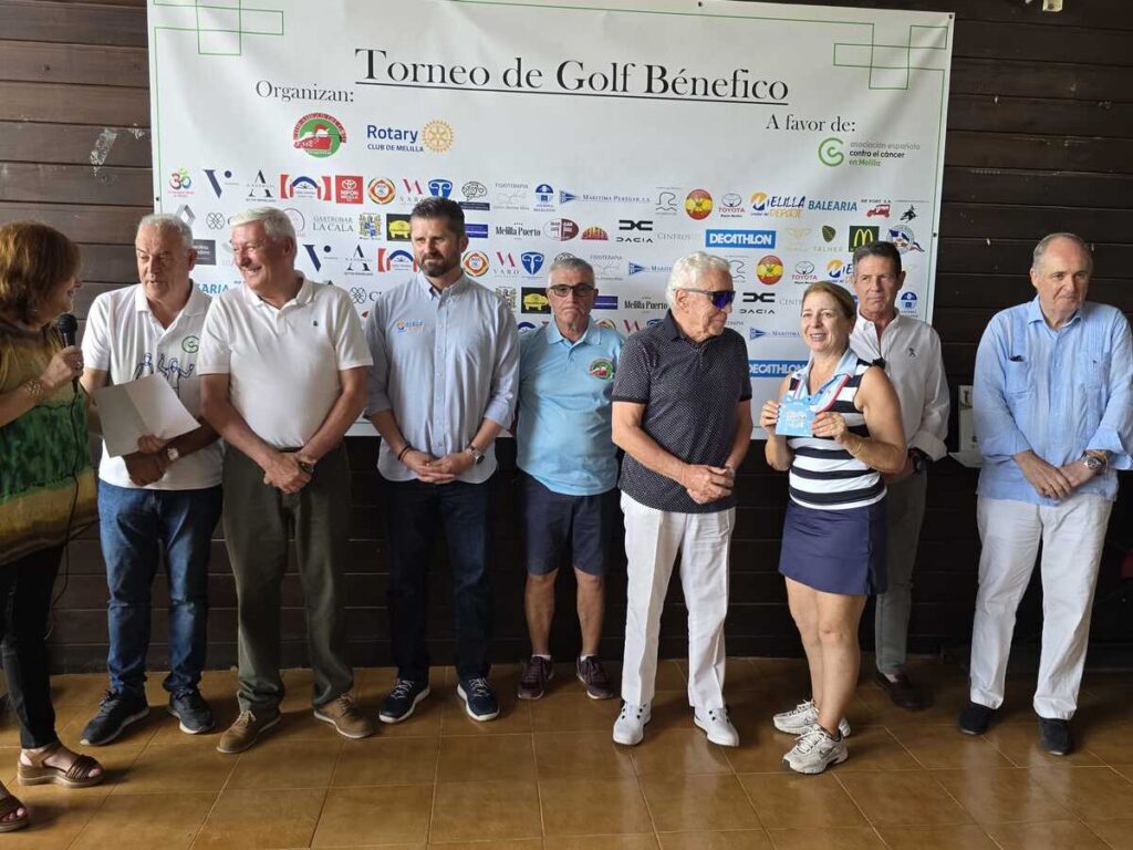 Group of people standing in a row at a charity golf tournament recognition event, with a banner reading 'Torneo de Golf Benefico' in the background (A favor de: ...). Women holds a microphone on the left while others smile and chat.
