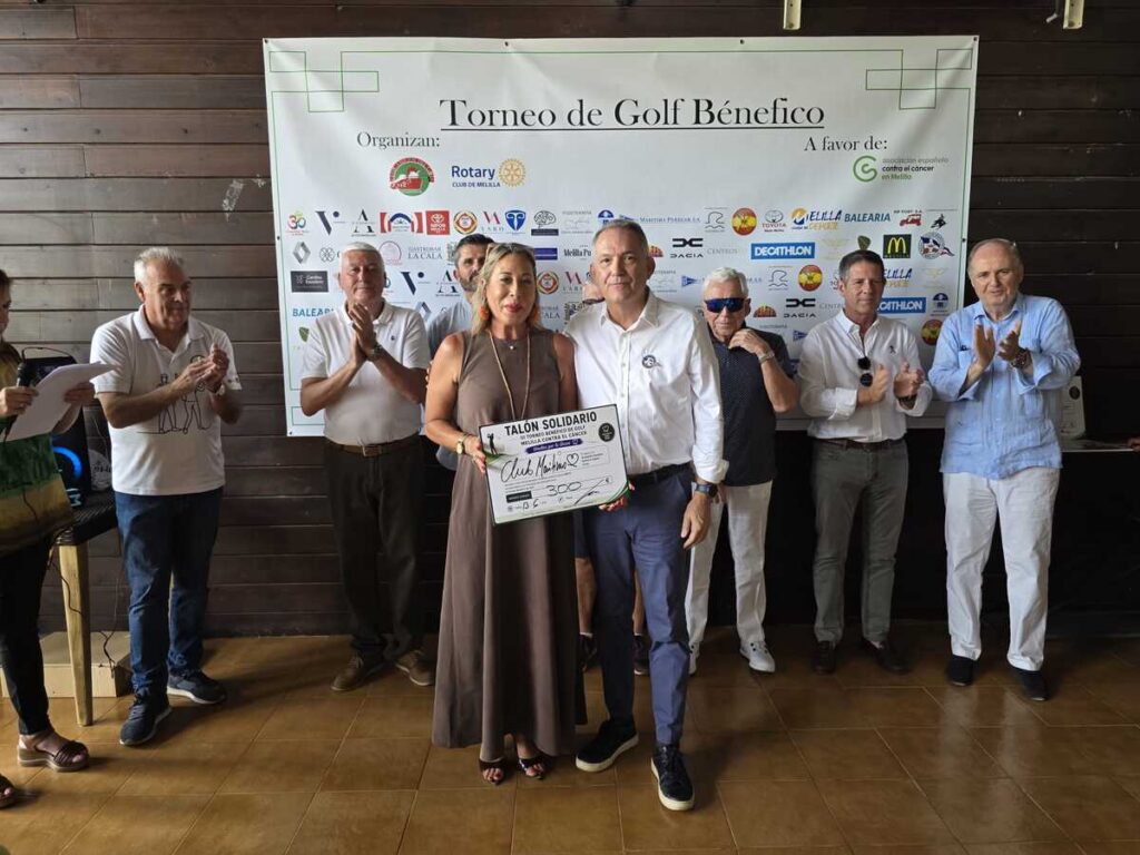 Group of people on a stage applauding at a charity golf tournament, with a banner reading 'Torneo de Golf Benéfico' behind them and a woman holding a large donation check.
