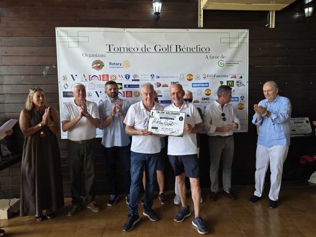 Group of people at a charity golf tournament presenting a large donation check while others applaud in front of a sponsor banner.