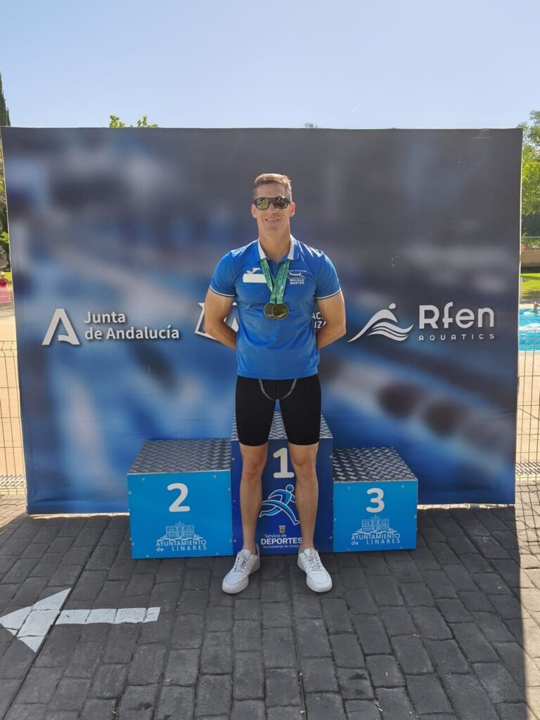 Male athlete on the first-place podium wearing a blue shirt, black shorts, sunglasses, and medals around his neck, with a blue sponsor backdrop (Junta de Andalucía / Rfen Aquatics).