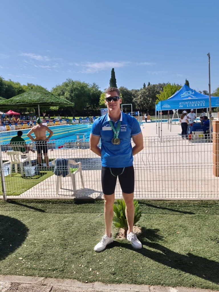 Man in a blue polo and black shorts with medals around his neck standing poolside at a swimming competition, with tents and spectators in the background.