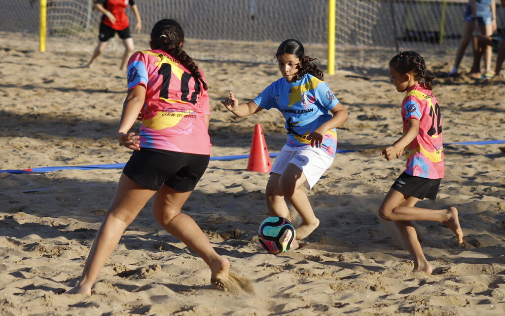 Girls playing beach soccer on sand, chasing a colorful ball near a blue-and-pink line marking with cones in the background. Two players in pink jerseys flank a player in blue, all focused on the ball.