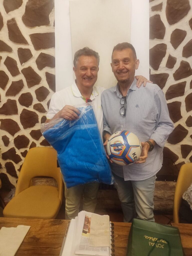 Dos hombres sonrientes sosteniendo un balón de fútbol y una camiseta en un homenaje