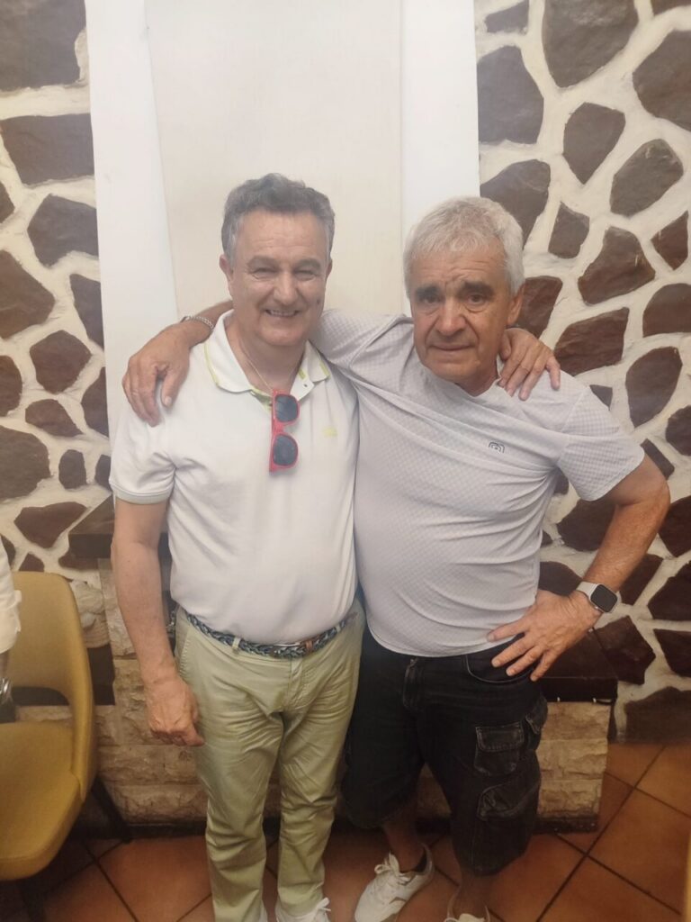 Two smiling men with arms around each other pose for a photo indoors against a stone wall.