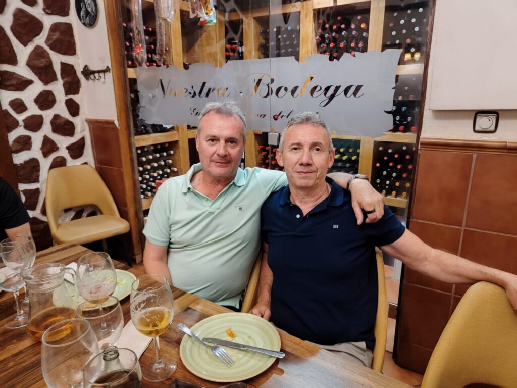 Two middle‑aged men sit close together at a restaurant table, one with his arm around the other, wine bottles on display behind them.