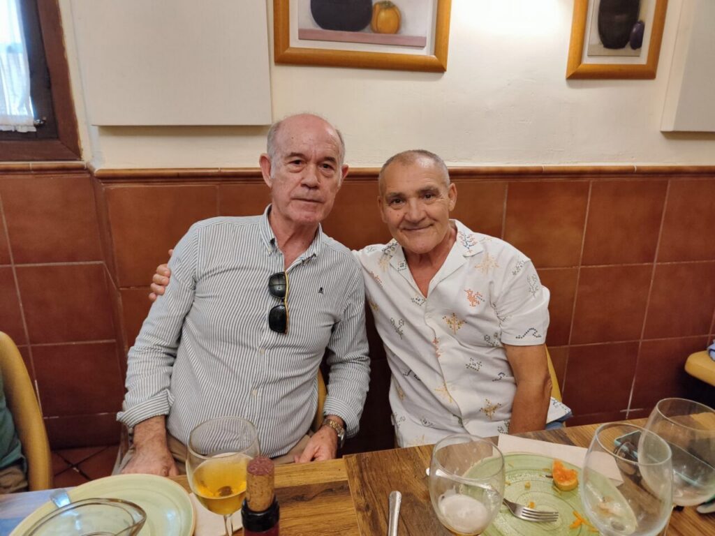 Two older men sit close together at a restaurant table, one with sunglasses hanging from his striped shirt, smiling at the camera.