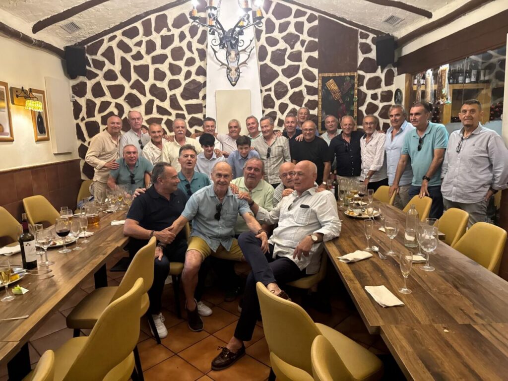 Group of men at a long wooden table in a rustic restaurant, posing and smiling for a group photo.