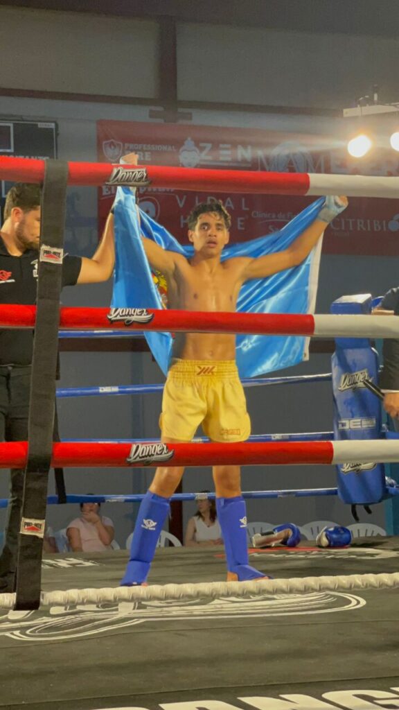 Young male boxer in yellow shorts and blue shin guards raises arms in victory inside a boxing ring, holding a blue cloth behind him.