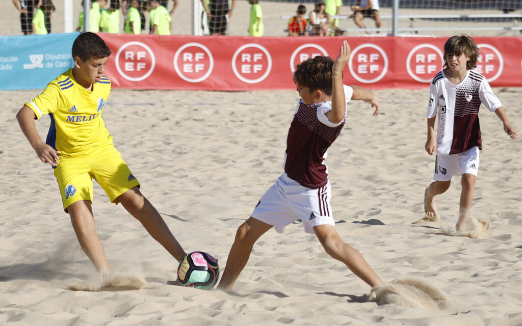 Three boys play beach soccer on sand; a yellow-clad player challenges two maroon-and-white teammates for the ball.