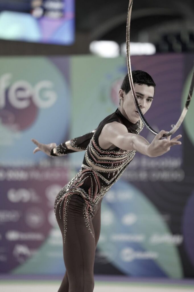 Male rhythmic gymnast in a jeweled maroon leotard performing with a hoop on a competition floor.