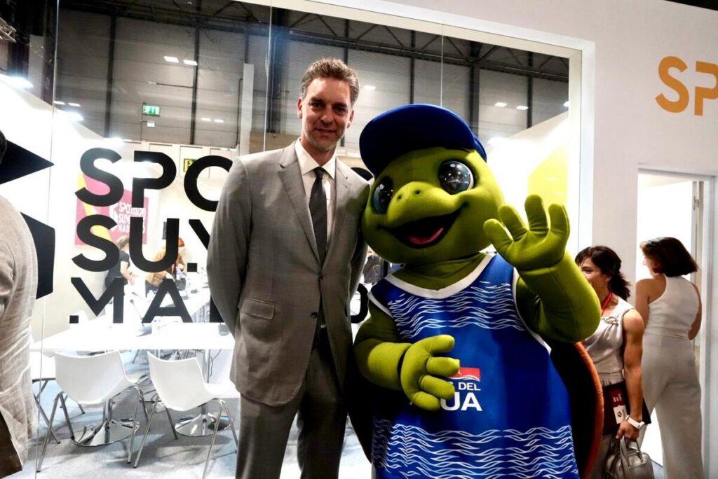 Man in a gray suit posing with a friendly green turtle mascot in a blue jersey at a trade show booth