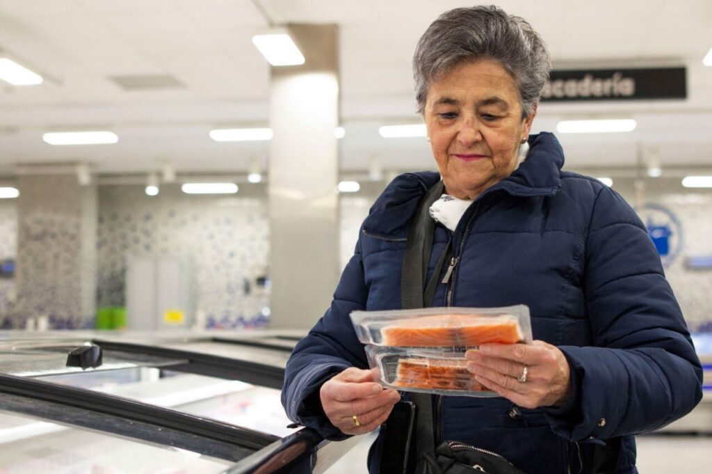 Older woman in a navy jacket holds two packages of salmon at a supermarket deli counter.