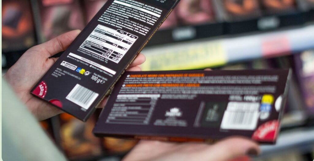 Person holds two dark purple chocolate boxes in a store, showing nutrition labels on the back within view of a shopper's hands.