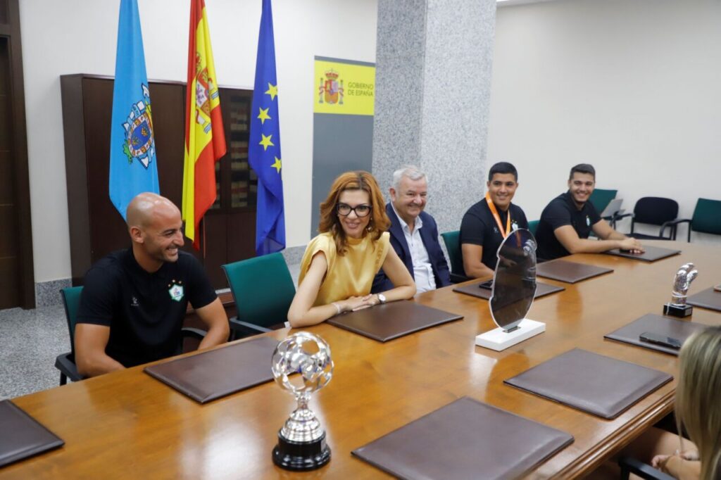 Five people sit at a wood conference table with folders; Spain and EU flags behind them, glass trophies on the table ahead of the group.
