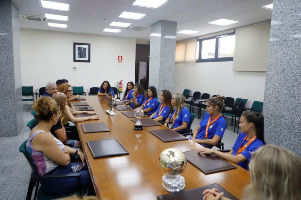 Group of people seated around a long conference table in a bright meeting room; several wear blue shirts with orange lanyards and face a panel, with trophies on the table.