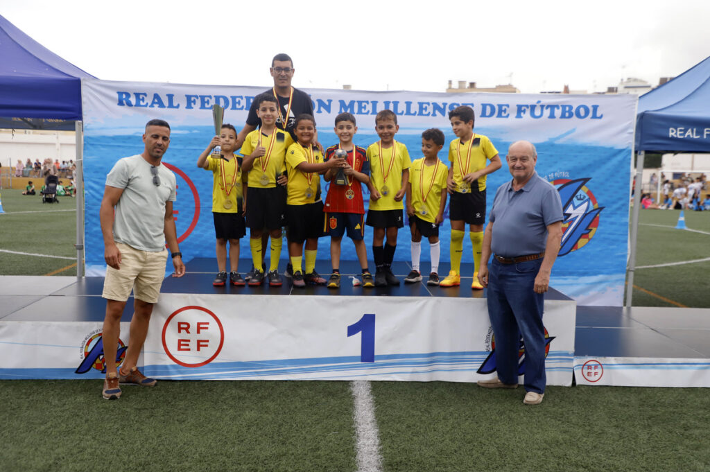 Group of young soccer players in yellow uniforms on a winners' podium, holding trophies and medals, with two adults flanking them on an outdoor field.