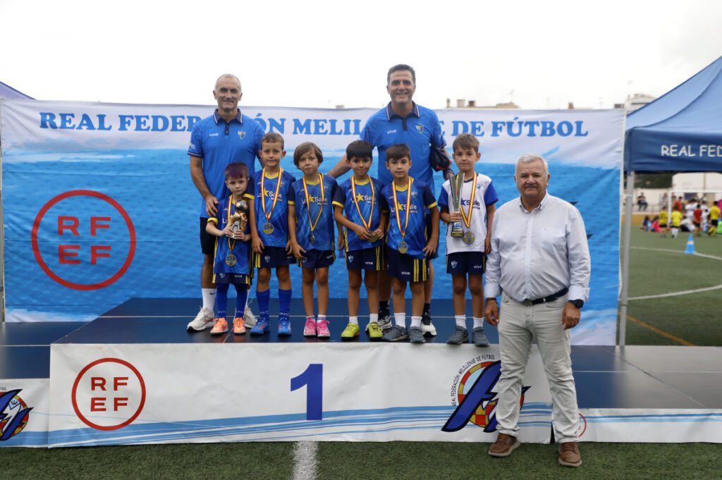 Group of young soccer players on a first-place podium wearing blue uniforms with medals, flanked by two coaches and an official in a light shirt far right, at a Real Federación Melilla de Fútbol event.