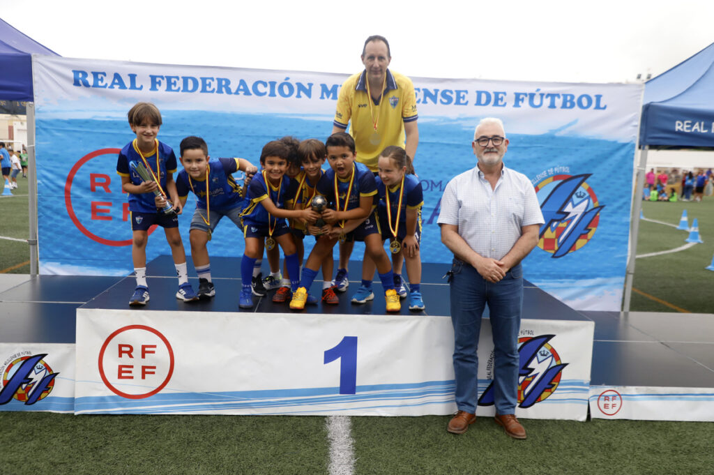 Young boys in blue team uniforms on a winner's podium, receiving medals and a trophy with two adults nearby on a football field.