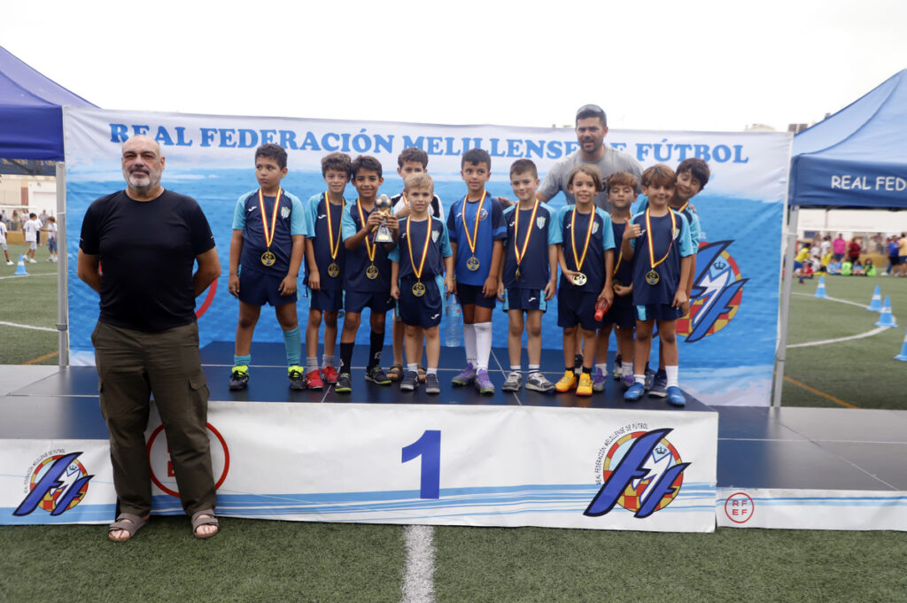 Group of young soccer players with medals standing on a winner's podium, flanked by two coaches, at a sports event.