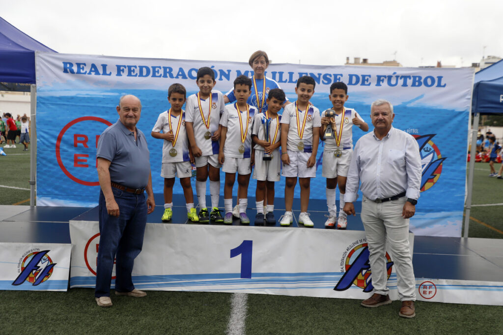 Group of young soccer players on a first-place podium wearing white uniforms and medals, flanked by two men, at an outdoor awards ceremony with a blue banner backdrop.