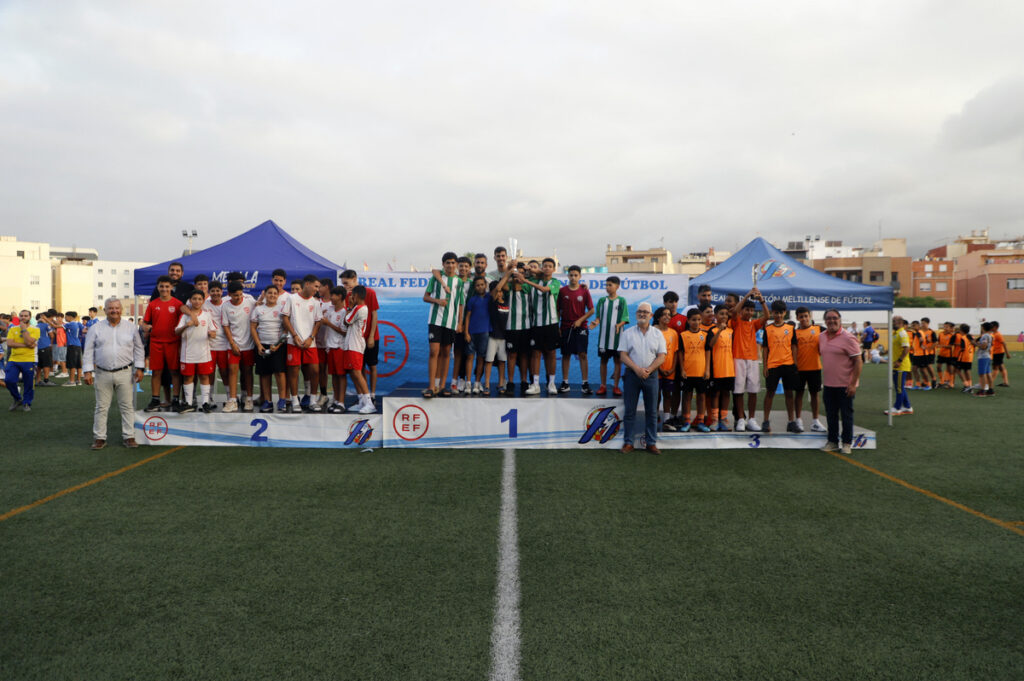 Youth soccer teams pose on a tiered podium after a tournament awards ceremony on a green field with blue tents in the background.