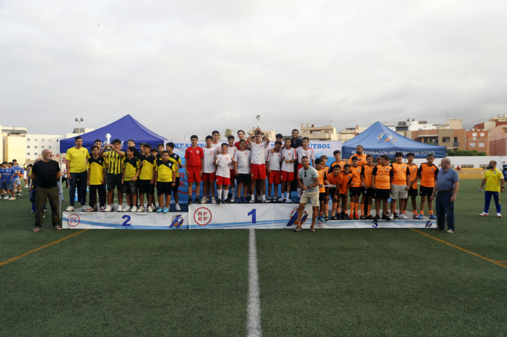 Youth soccer teams stand on a three-tier podium on a football field, celebrating with a trophy as tents loom behind them.