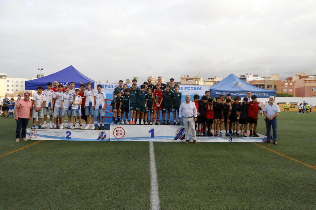Youth soccer awards ceremony on a turf field with a three-tier podium and players in white, green, and red uniforms posing with coaches and organizers.