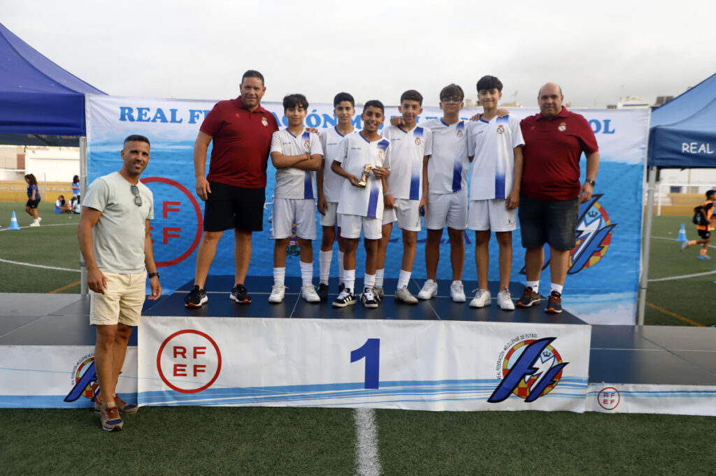 Group of young soccer players in white uniforms on a winner's podium holding a trophy, flanked by two men in red shirts, outdoors at a tournament.