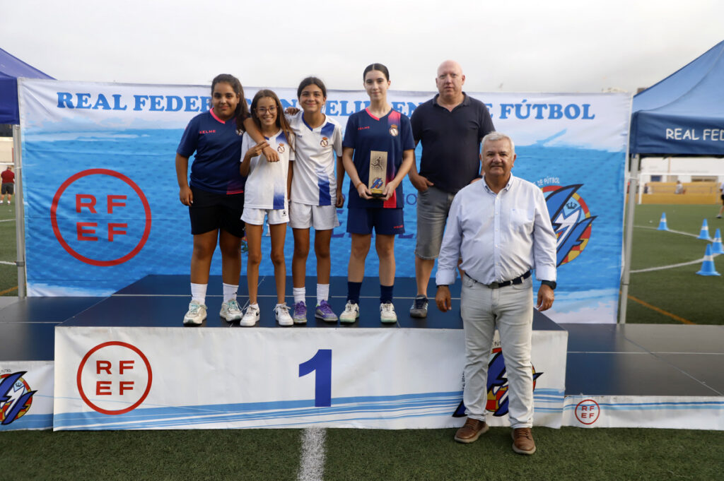 Group of young female soccer players on a 1st-place podium holding a trophy, flanked by two adults, with a Real Federación de Fútbol backdrop.