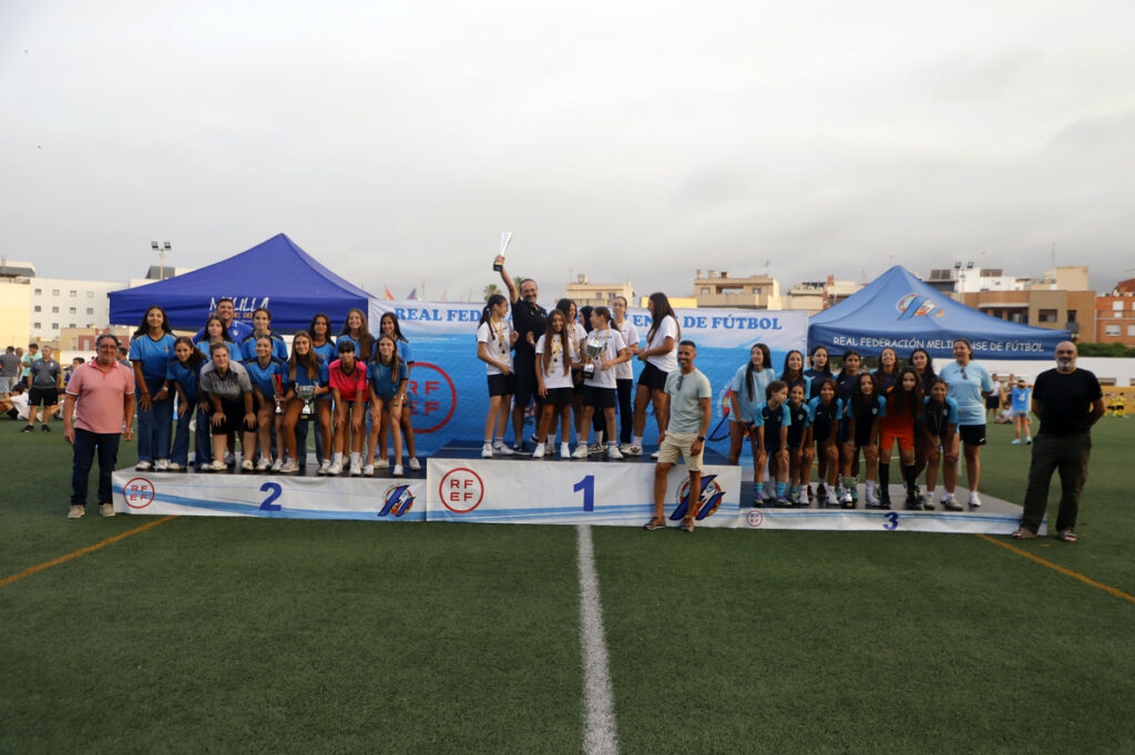 Group of young female players on a winners' podium (1st, 2nd, 3rd) at an outdoor soccer field, flanked by coaches and blue tents behind them.