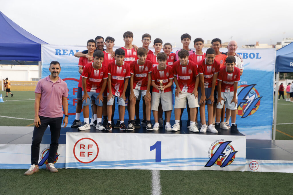 Youth soccer team in red jerseys stands on a first-place podium holding a trophy, celebrating win with coaches nearby.
