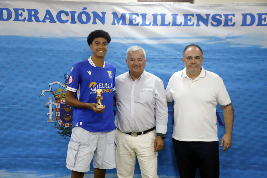 Young soccer player in blue jersey holding a trophy, flanked by two men in white shirts at an awards event.