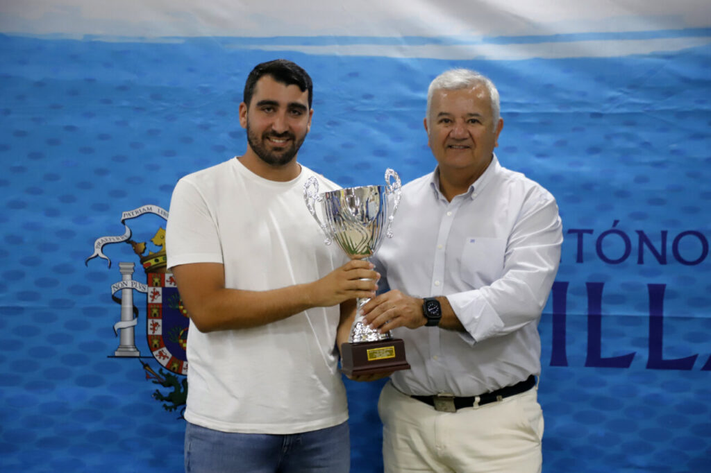 Two men hold a large silver trophy together at an award ceremony, standing before a blue patterned backdrop and crest.